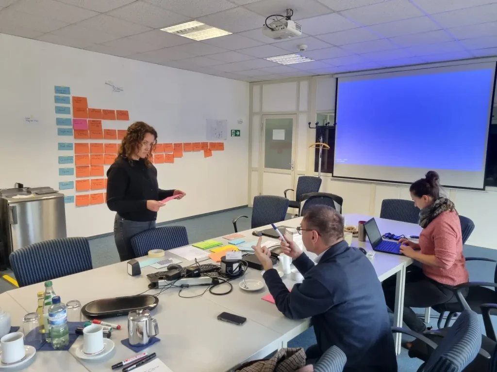 Drei Personen gruppieren sich um einen Tisch. Im Hintergrund hängen Post-Its an der Wand und eine Leinwand